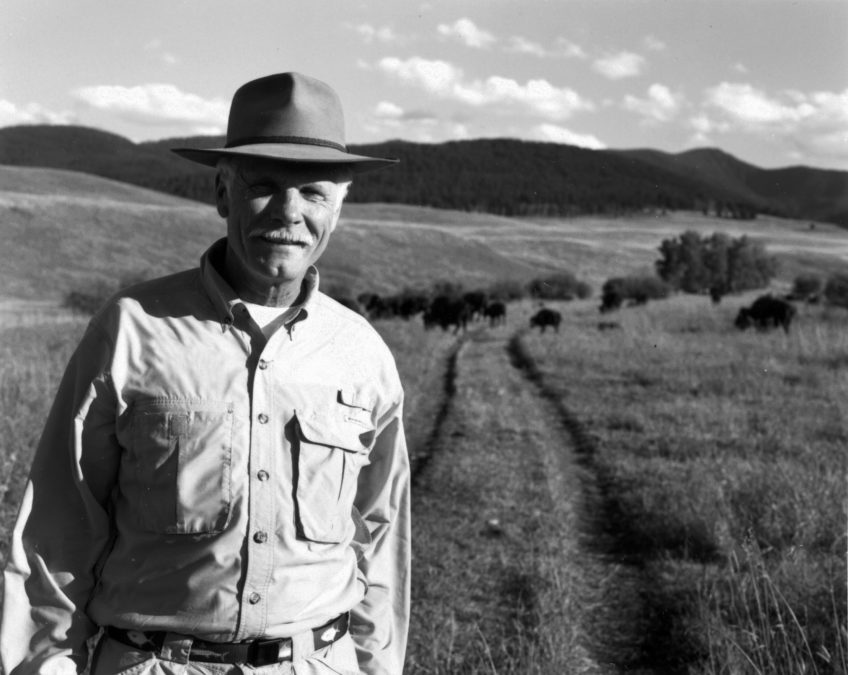 a black and white photo of Ted Turner, who stands on a wide plain with bison and mountains in the distance.